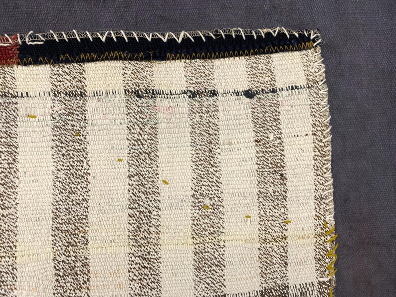 Puede incluir: Primer plano de una secci&oacute;n de un textil tejido con un patr&oacute;n de rayas en crema y marr&oacute;n. El borde est&aacute; acabado con un borde negro y costuras blancas. La esquina tiene un acento rojo y negro.