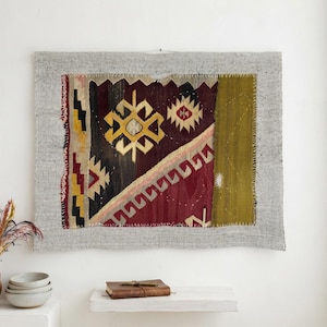 Op de afbeelding: Een rechthoekig wandkleed met een centraal paneel van donkerbordeaux, zwarte en gele geometrische patronen, genaaid op een lichtgrijze, linnenachtige stoffen rand. Het textiel vertoont zichtbare handgestikte randen en een verticaal olijfgroen gedeelte.