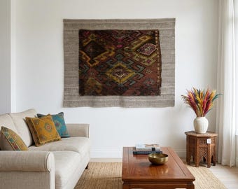 May include: A living room scene featuring a beige sofa with decorative pillows in shades of gold, teal, and patterned designs. A wooden coffee table holds books and a brass bowl. A large, patterned rug hangs on the wall above the sofa.