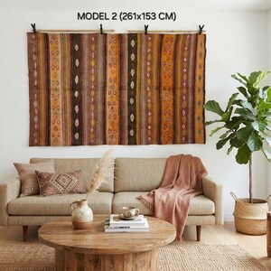 Op de afbeelding: Een groot rechthoekig wandkleed met het label MODEL 2 (261x153 cm) met verticale strepen in bruin-, oranje- en geeltinten met kleine ruitpatronen. De stof is opgehangen aan twee houten roedes met zwarte clips.