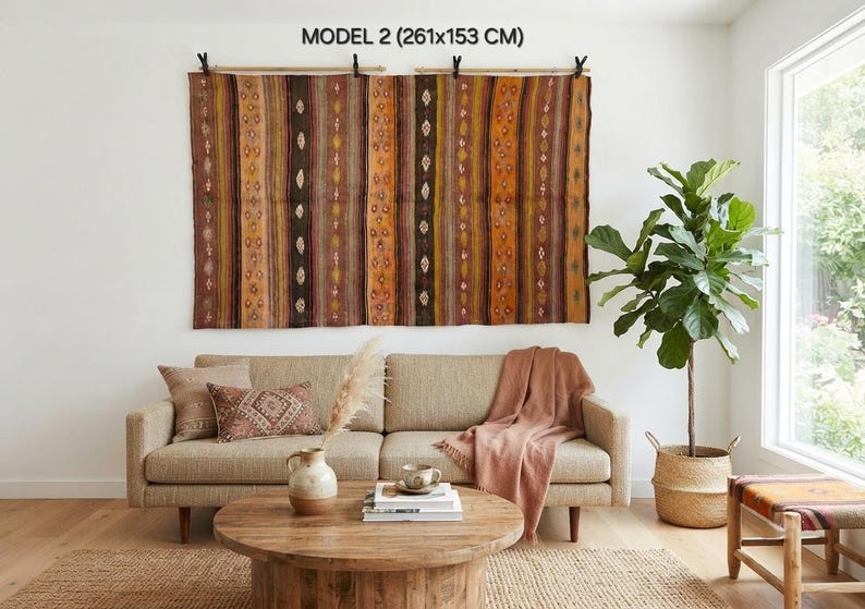 Op de afbeelding: Een groot rechthoekig wandkleed met het label MODEL 2 (261x153 cm) met verticale strepen in bruin-, oranje- en geeltinten met kleine ruitpatronen. De stof is opgehangen aan twee houten roedes met zwarte clips.