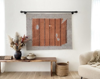Puede incluir: Un tapiz rectangular con un panel central de color naranja óxido con rayas verticales de color marrón oscuro, rodeado por un borde gris. El tejido muestra costuras visibles y bordes deshilachados, suspendido de una barra de metal negra.