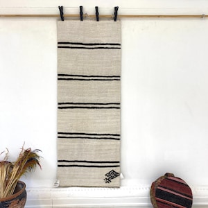 May include: A beige and black woven rug with four horizontal black stripes. The rug is hanging from a wooden rod with black clips. A decorative pattern is at the bottom right corner.