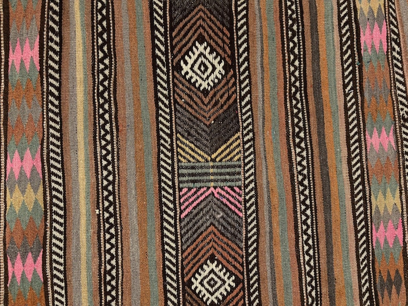 Op de afbeelding: Een geweven textiel met verticale strepen in tinten bruin, beige, grijs en groenblauw. Het patroon bevat herhalende geometrische ruitvormen in roze, geel en groen, gescheiden door smalle zwart-witte zigzaglijnen als randen.