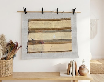 May include: A rectangular woven textile wall hanging featuring horizontal stripes in shades of cream, tan, and dark brown. The fabric has a frayed edge on the left side and is suspended from a wooden rod by four black metal clips.