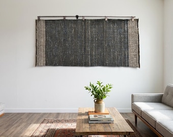 May include: A large rectangular wall hanging featuring a dark charcoal gray woven fabric with textured light brown vertical borders on each side, suspended from a horizontal metal rod with silver clips attached to the top edge of the textile.