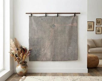 Puede incluir: Un tapiz rectangular grande de tela gris suspendido de una barra de madera oscura. El tejido presenta sutiles líneas punteadas finas que forman un patrón similar a un mapa en su superficie, con costuras visibles a lo largo de los bordes.