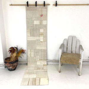 Pode incluir: Um tapete comprido, retangular, de cor branco-queimado feito de tecido patchwork. O tapete está pendurado numa parede com uma vara de madeira. Uma cadeira branca com um assento de tecido tecido está em primeiro plano.