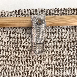 Puede incluir: Primer plano de una tela tejida con una barra de madera. La tela es una mezcla de hilos marrones y crema, con un aspecto texturizado. Un lazo de tela con un ojal met&aacute;lico asegura la tela a la barra.