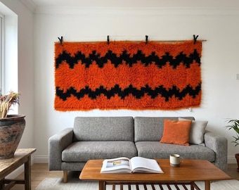 May include: An orange and black zig-zag patterned wall hanging. The shag rug is mounted on a wooden rod with black clips. The rug hangs above a gray sofa with orange and patterned pillows. A wooden coffee table and a large terracotta pot are in the foreground.