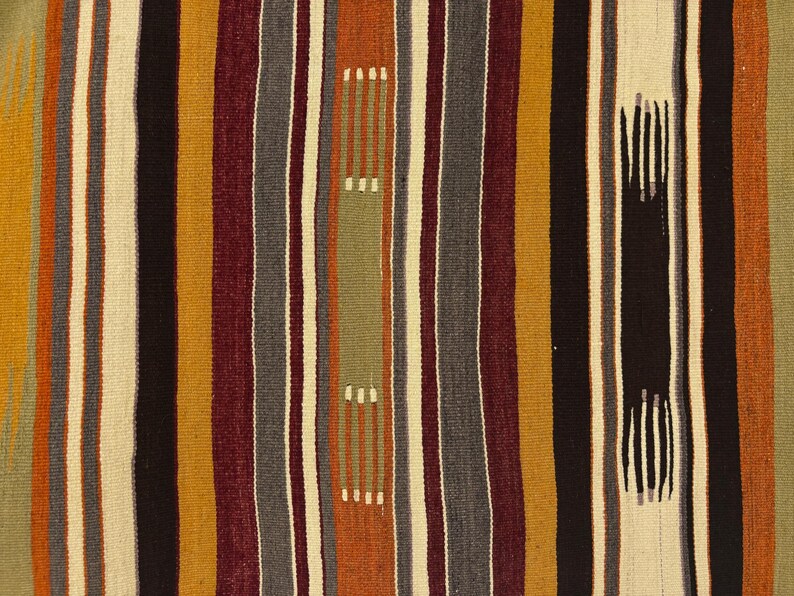 May include: A woven textile featuring vertical stripes in shades of mustard yellow, deep burgundy, charcoal gray, cream, black, and muted sage green. The pattern includes intermittent dashed lines and solid blocks of color throughout the fabric surface.