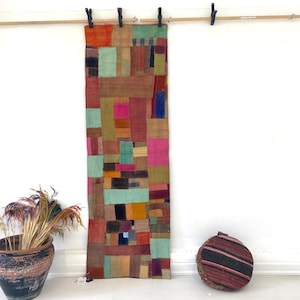 Pode incluir: Um têxtil colorido em patchwork pendurado numa vara de madeira com clipes pretos. O têxtil apresenta blocos retangulares de várias cores, incluindo rosa, verde e castanho. Um vaso decorativo com plantas secas e uma almofada redonda listrada estão no chão.