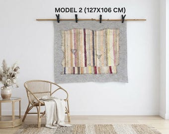 Puede incluir: Un tapiz rectangular con un panel central de rayas verticales multicolores en tonos rosa, amarillo y marrón, bordeado por una tela gris sólida. La pieza está suspendida de una barra de madera y mide 127x106 cm.