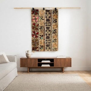 May include: A rectangular woven wall hanging featuring a patchwork of intricate patterns in shades of brown, beige, red, and black, suspended from a horizontal wooden rod mounted on a plain white wall above a low wooden media console cabinet.