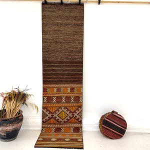 May include: A long, rectangular runner rug featuring a solid dark brown upper section that transitions into a lower section decorated with intricate geometric patterns in shades of orange, yellow, and white, finished with a fringed bottom edge.