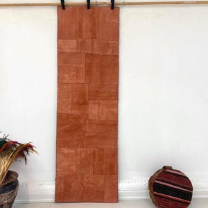Puede incluir: Una alfombra larga y rectangular de color óxido hecha de varios parches rectangulares. La alfombra cuelga de una barra de madera. Una maceta decorativa con plantas secas y un objeto de madera redondo y rayado están en el suelo.