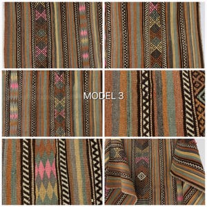 Op de afbeelding: Een geweven textiel met verticale strepen in tinten bruin, beige, blauw en groen, geaccentueerd door geometrische ruitpatronen in roze, geel en groenblauw. De stof toont een herhalende reeks van gestructureerde banden en de tekst MODEL 3.