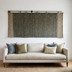 Op de afbeelding: Een rechthoekig geweven wandkleed met een donkergroen middenpaneel geflankeerd door twee beige verticale zijpanelen, opgehangen aan een zilveren metalen roede met clips. Het textiel heeft een grove, zichtbare textuur en hangt horizontaal.