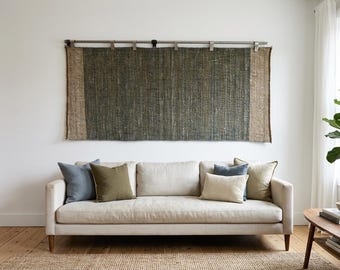 May include: A rectangular woven wall hanging featuring a dark green center panel flanked by two tan vertical side panels, suspended from a silver metal rod with clips. The textile has a visible coarse texture and hangs horizontally above a sofa.