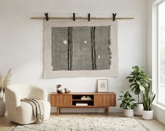 Puede incluir: Un tapiz rectangular con un panel central de color verde salvia, dos franjas verticales negras y bordes deshilachados, suspendido de una varilla de madera mediante cuatro clips negros. El tejido presenta una textura con detalles blancos.