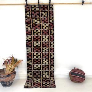 May include: A long, rectangular runner rug featuring a dark black base with a repeating grid pattern. Each square contains a geometric cross-like motif in alternating shades of cream, yellow, and deep red, finished with a short fringe at the top.