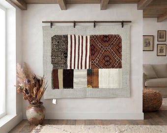 May include: A rectangular wall hanging tapestry featuring a patchwork design of various woven fabric segments in shades of brown, cream, black, and white, suspended from a dark wooden rod mounted horizontally on a light-coloured wall.