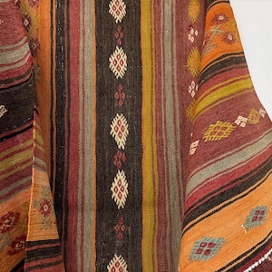 Op de afbeelding: Een geweven textiel met verticale strepen in tinten oranje, rood, bruin en geel. De stof is versierd met herhalende ruitvormige geometrische patronen in wit, rood en blauw, gerangschikt in kolommen over het getextureerde oppervlak.