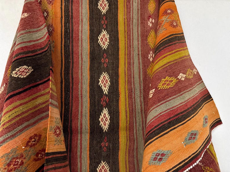 Op de afbeelding: Een geweven textiel met verticale strepen in tinten oranje, rood, bruin en geel. De stof is versierd met herhalende ruitvormige geometrische patronen in wit, rood en blauw, gerangschikt in kolommen over het getextureerde oppervlak.