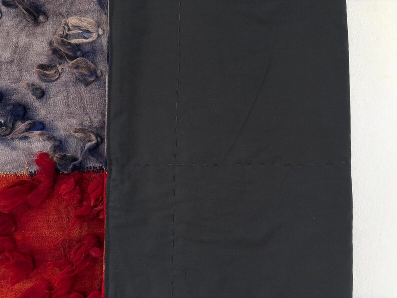 May include: A textile artwork featuring a patchwork design. The left side showcases a blend of textures and colors, including red, orange, gray, and blue. The right side is a solid black fabric. The piece is likely a wall hanging or decorative textile.
