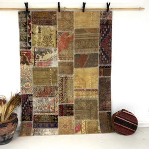 May include: A patchwork rug with a variety of colors and patterns, including beige, brown, red, and green. The rug is hanging from a wooden rod with black clips. A decorative pot with dried plants and a striped round pillow are on the floor.