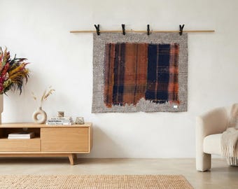 May include: A rectangular wall hanging featuring a central panel of brown and navy blue plaid fabric with frayed edges, bordered by a textured gray woven frame. The piece is suspended from a wooden rod by four black clips against a plain wall.