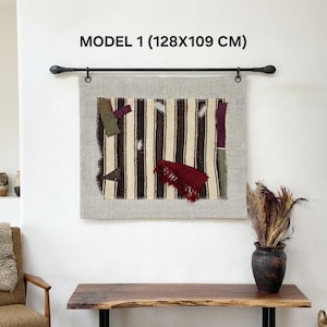 Puede incluir: Un tapiz rectangular con un panel de tela a rayas marrones y crema con parches en verde oliva, morado y rojo. La pieza está suspendida de una barra de metal negro y mide 128x109 cm, tal como indica el texto MODEL 1.