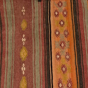 Op de afbeelding: Een geweven textielkleed met verticale strepen in tinten donkerbruin, gedempt rood, saliegroen en oranje. Het ontwerp bevat herhalende geometrische ruitmotieven in cr&egrave;me, geel en bordeauxrood, gerangschikt in verticale kolommen over de stof.