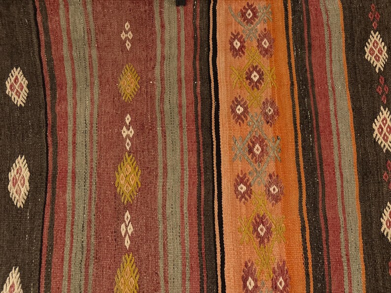 Op de afbeelding: Een geweven textielkleed met verticale strepen in tinten donkerbruin, gedempt rood, saliegroen en oranje. Het ontwerp bevat herhalende geometrische ruitmotieven in cr&egrave;me, geel en bordeauxrood, gerangschikt in verticale kolommen over de stof.