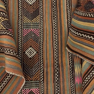 Op de afbeelding: Een geweven textiel met een herhalend patroon van verticale strepen en geometrische vormen in tinten bruin, beige, gedempt groen en roze accenten. De stof vertoont een geweven textuur met ingewikkelde zigzag- en ruitmotieven door het hele ontwerp.