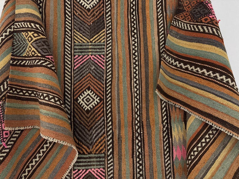 Op de afbeelding: Een geweven textiel met een herhalend patroon van verticale strepen en geometrische vormen in tinten bruin, beige, gedempt groen en roze accenten. De stof vertoont een geweven textuur met ingewikkelde zigzag- en ruitmotieven door het hele ontwerp.