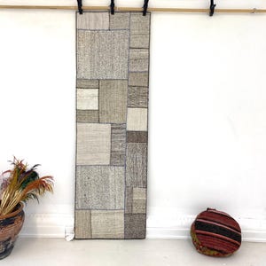 May include: A rectangular wall hanging rug featuring a patchwork design of various neutral-toned fabric squares in shades of beige, gray, and off-white. The rug is suspended from a wooden rod by black clips and displays a textured, woven surface.
