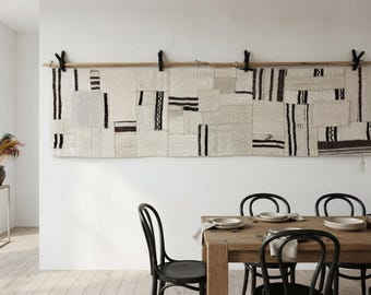 May include: A long, rectangular wall hanging composed of stitched-together off-white and cream fabric patches featuring black striped and geometric patterns. The textile is suspended from a horizontal wooden rod by four black metal clips.