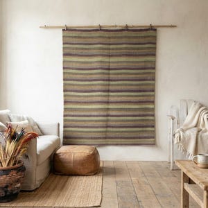 May include: A rectangular wall hanging featuring horizontal stripes in shades of olive green, brown, and muted purple. The textile is suspended from a thin wooden rod mounted horizontally on the wall, displaying a consistent woven texture throughout.