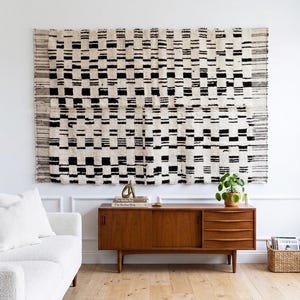 Peut inclure: Une grande tenture murale rectangulaire présentant un motif géométrique répétitif de bandes et de blocs tissés noirs et crème. Le textile présente une surface texturée avec des bords frangés sur les côtés gauche et droit, monté horizontalement.