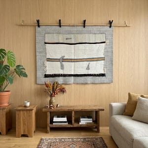 Puede incluir: Un salón de tonos neutros con un tapiz de pared tejido, un banco de madera y una planta en maceta. El tapiz rectangular tiene un borde gris y un centro crema con rayas negras y doradas. Un sofá beige y una alfombra estampada completan el espacio.
