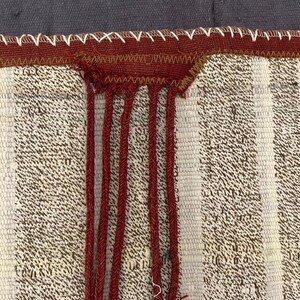 Puede incluir: Primer plano de un textil tejido con rayas verticales en crema y marr&oacute;n. Una secci&oacute;n de tela roja est&aacute; cosida al borde superior con hilo blanco. Cordones rojos cuelgan de la tela roja, agregando un elemento textural.