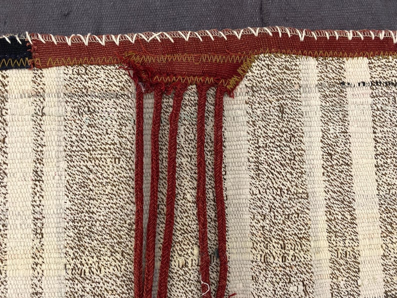 Puede incluir: Primer plano de un textil tejido con rayas verticales en crema y marr&oacute;n. Una secci&oacute;n de tela roja est&aacute; cosida al borde superior con hilo blanco. Cordones rojos cuelgan de la tela roja, agregando un elemento textural.