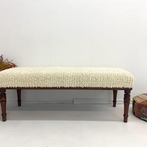 May include: A cream-colored upholstered bench with a textured surface and dark brown wooden legs. The bench is rectangular and has a low profile. The legs are turned and add a touch of elegance to the piece. A decorative item for the living room.