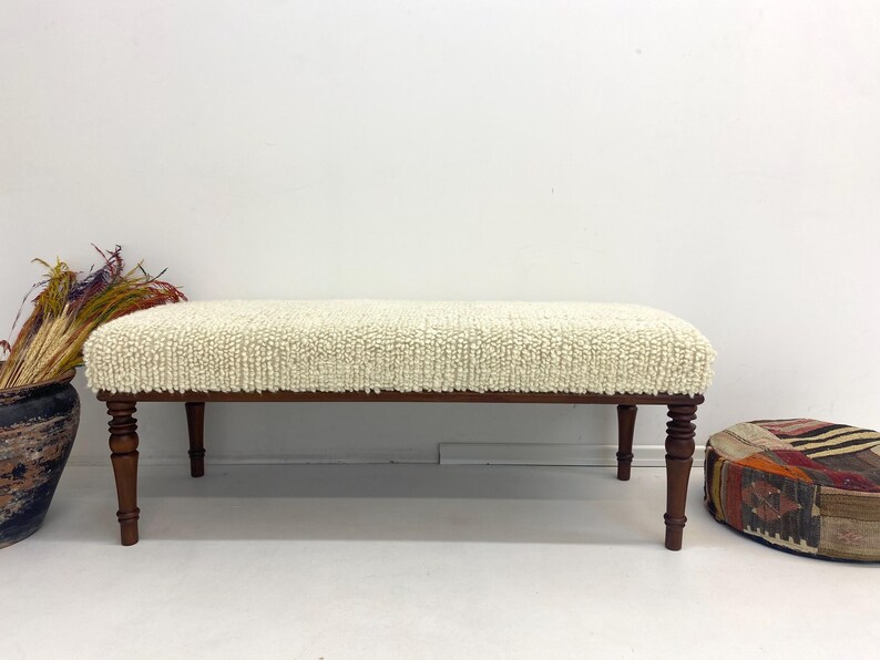 May include: A cream-colored upholstered bench with a textured surface and dark brown wooden legs. The bench is rectangular and has a low profile. The legs are turned and add a touch of elegance to the piece. A decorative item for the living room.
