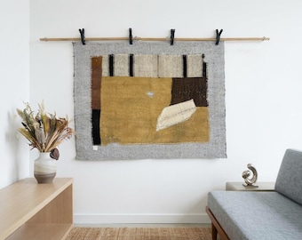 May include: A rectangular wall hanging featuring a gray border and a central panel of mustard yellow, brown, and cream fabric patches. The textile is suspended from a wooden rod by four black clips, displaying a mix of textures and geometric shapes.