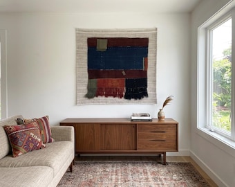 May include: A woven wall hanging with a rectangular design in shades of blue, red, and brown, with a cream-colored border. The wall hanging is displayed above a wooden cabinet in a living room setting.