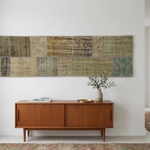 May include: A long, rectangular patchwork wall hanging featuring muted earth tones, olive greens, and faded blues in a grid pattern. The textile displays distressed textures and subtle geometric motifs, mounted horizontally above a wooden sideboard.
