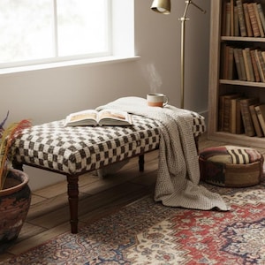 May include: A comfortable interior scene with a bench featuring a brown and cream checkered pattern, a blanket, and an open book. A steaming mug sits on the bench. A terracotta pot holds dried plants. A bookshelf and patterned rug complete the scene.