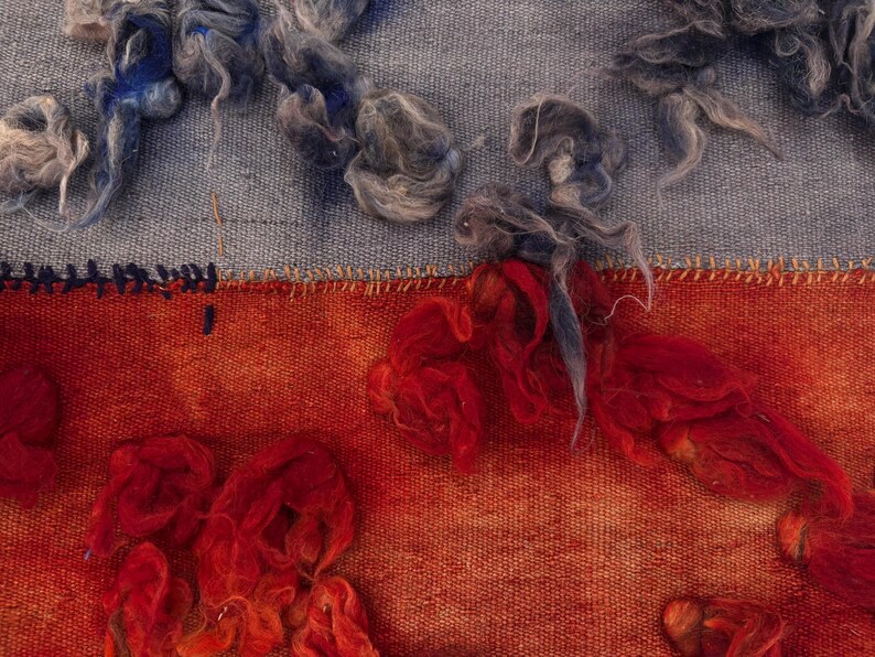 May include: Close-up of a textile artwork featuring a blend of textures and colors. The upper section displays a light blue fabric with tufts of gray and blue fibers. The lower section showcases a vibrant orange-red hue with similar fiber accents.